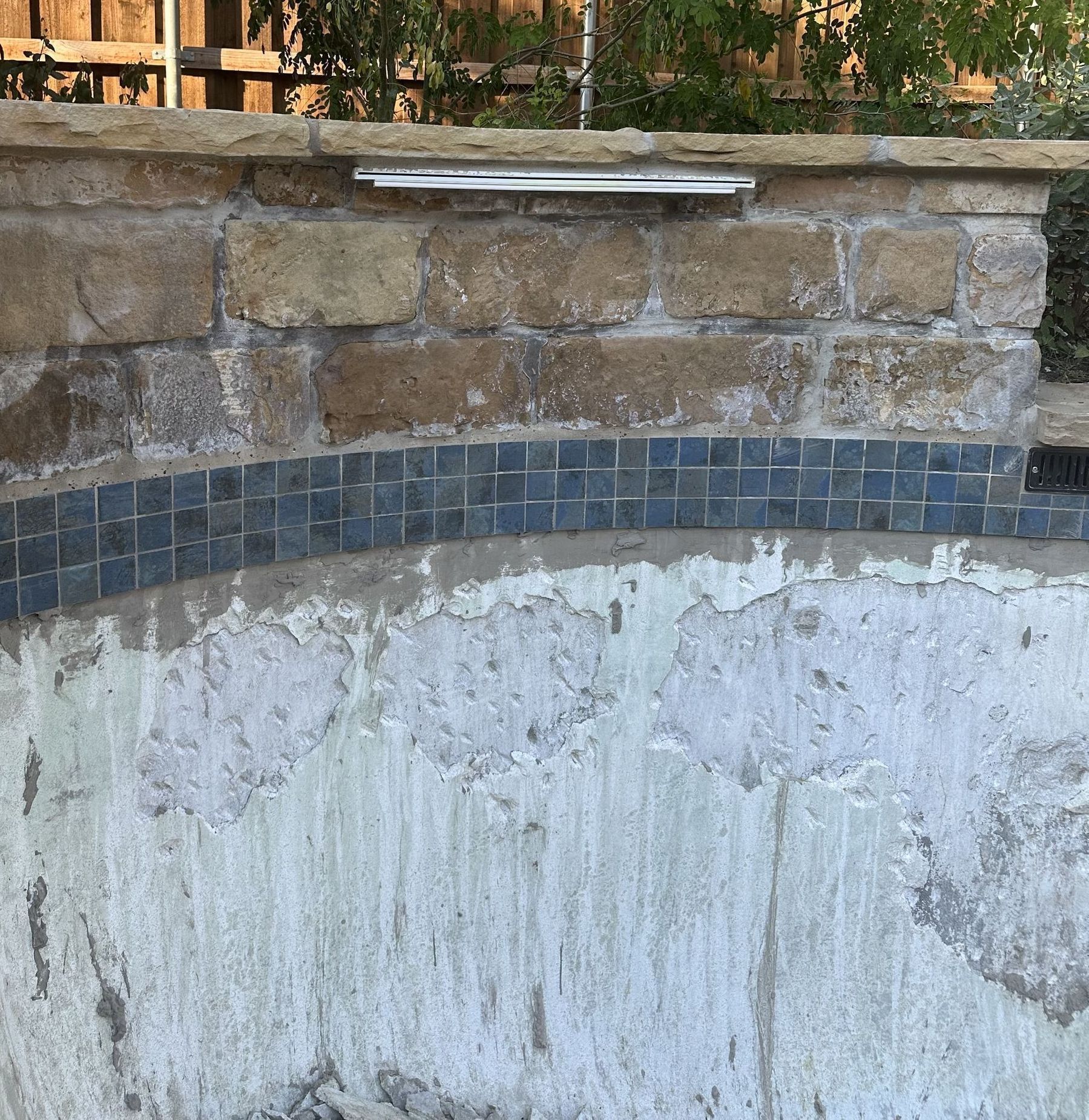 Pool wall showing damaged plaster below blue tile border and stone wall above.