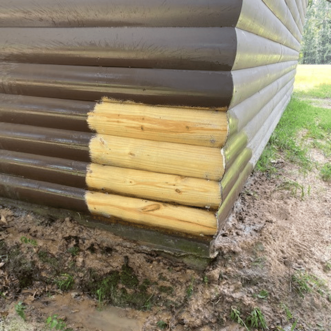 A partially painted brown log cabin corner; fresh paint contrasts with unpainted wood, muddy ground, and grass.