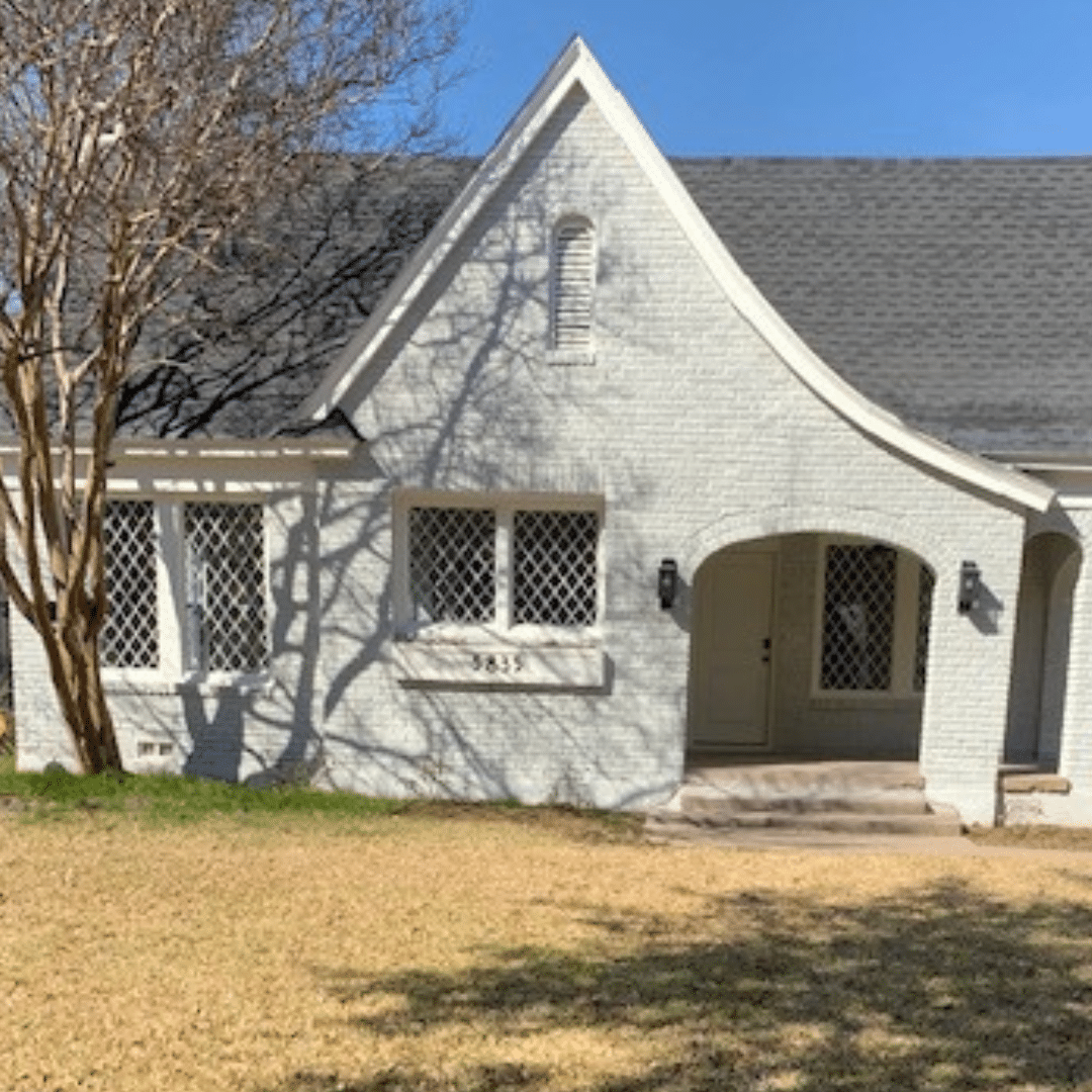 White-painted Tudor-style house with arched entryway and diamond-paned windows; bare tree casts shadows on the facade.