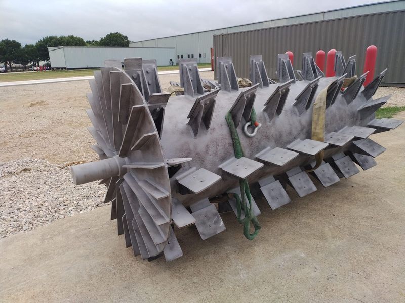 Large metal turbine-like industrial part resting on concrete, with blades and central shaft.