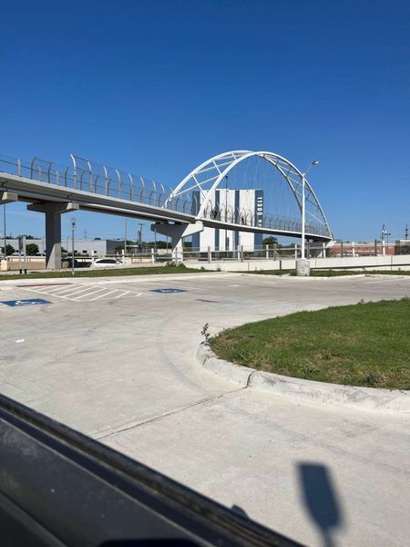Pedestrian bridge with white arch over a road and parking area on a sunny day.