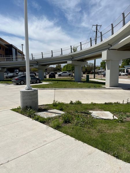 Elevated train tracks above a concrete area with a pole and some grass.