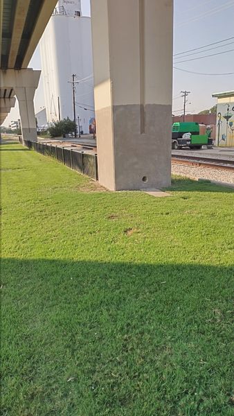 Green grass under a bridge with train tracks and a green truck passing by.