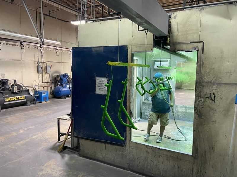 A person spray paints green bicycle frames inside a paint booth. Industrial setting with machinery and compressed air.