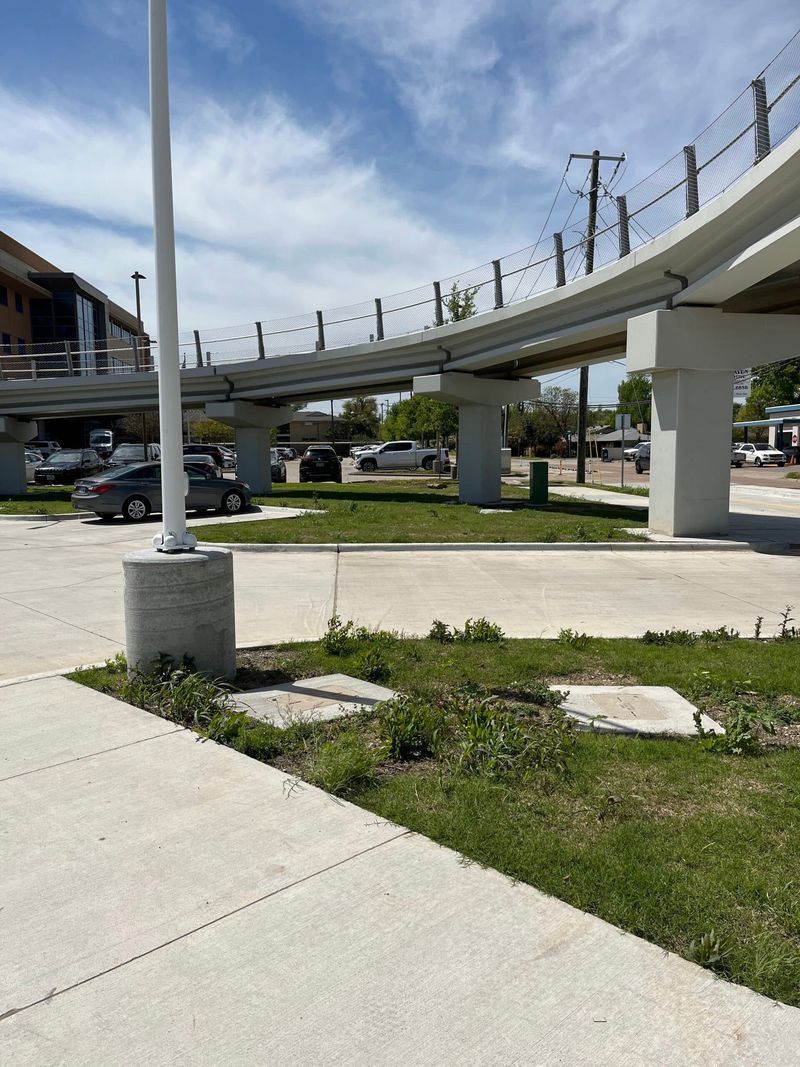 Elevated train tracks over a paved area and grassy patches. Blue sky with clouds.