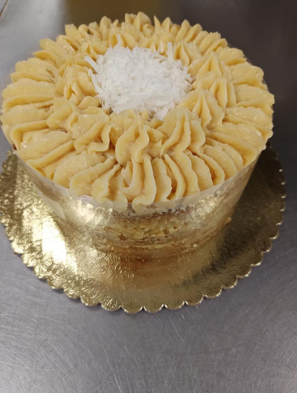 A round cake on a gold plate, topped with piped tan frosting and a center mound of white coconut flakes.