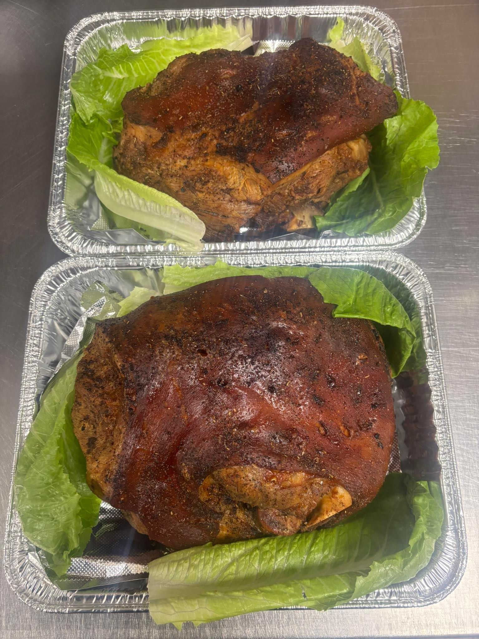 Two foil containers, each holding a seasoned, roasted pork knuckle resting on fresh green lettuce leaves.