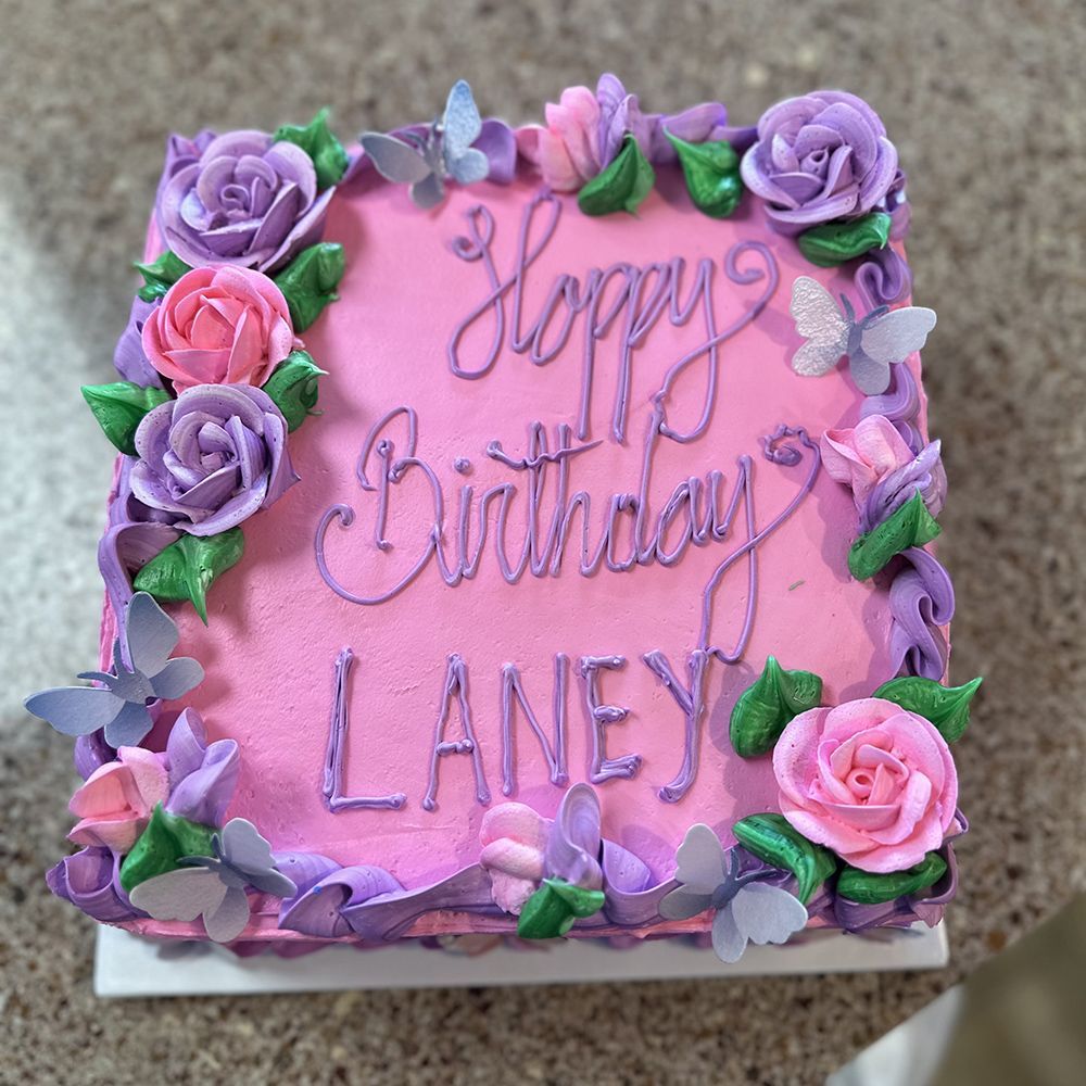 Pink square cake decorated with purple and pink frosting roses, green leaves, and butterflies; 