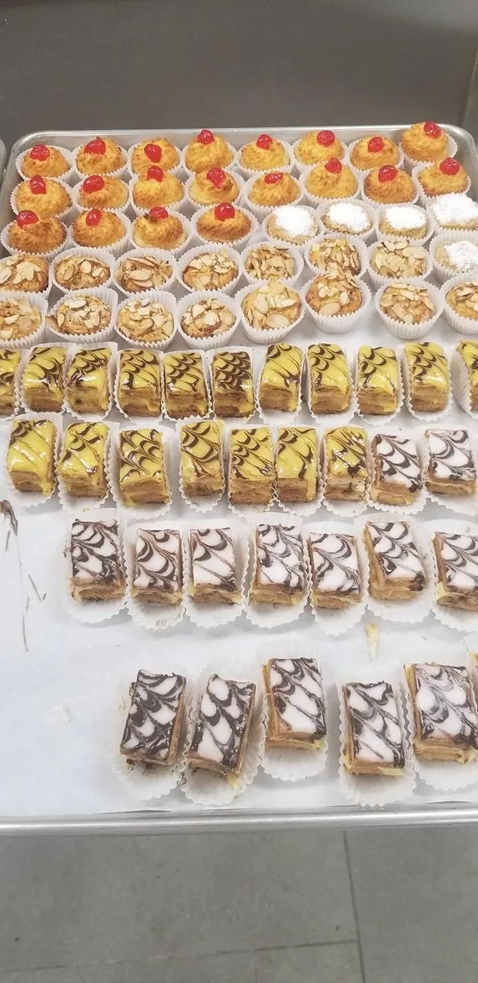 Tray of various pastries: mini cakes, tarts, and cherry-topped treats arranged on a reflective surface.