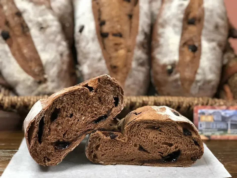 Sliced chocolate bread with olive inclusions, bread loaves in basket in background.