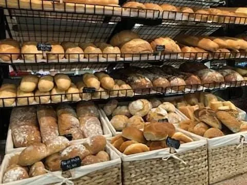 Shelves of bread at a bakery, various loaves and rolls displayed in baskets and on racks.