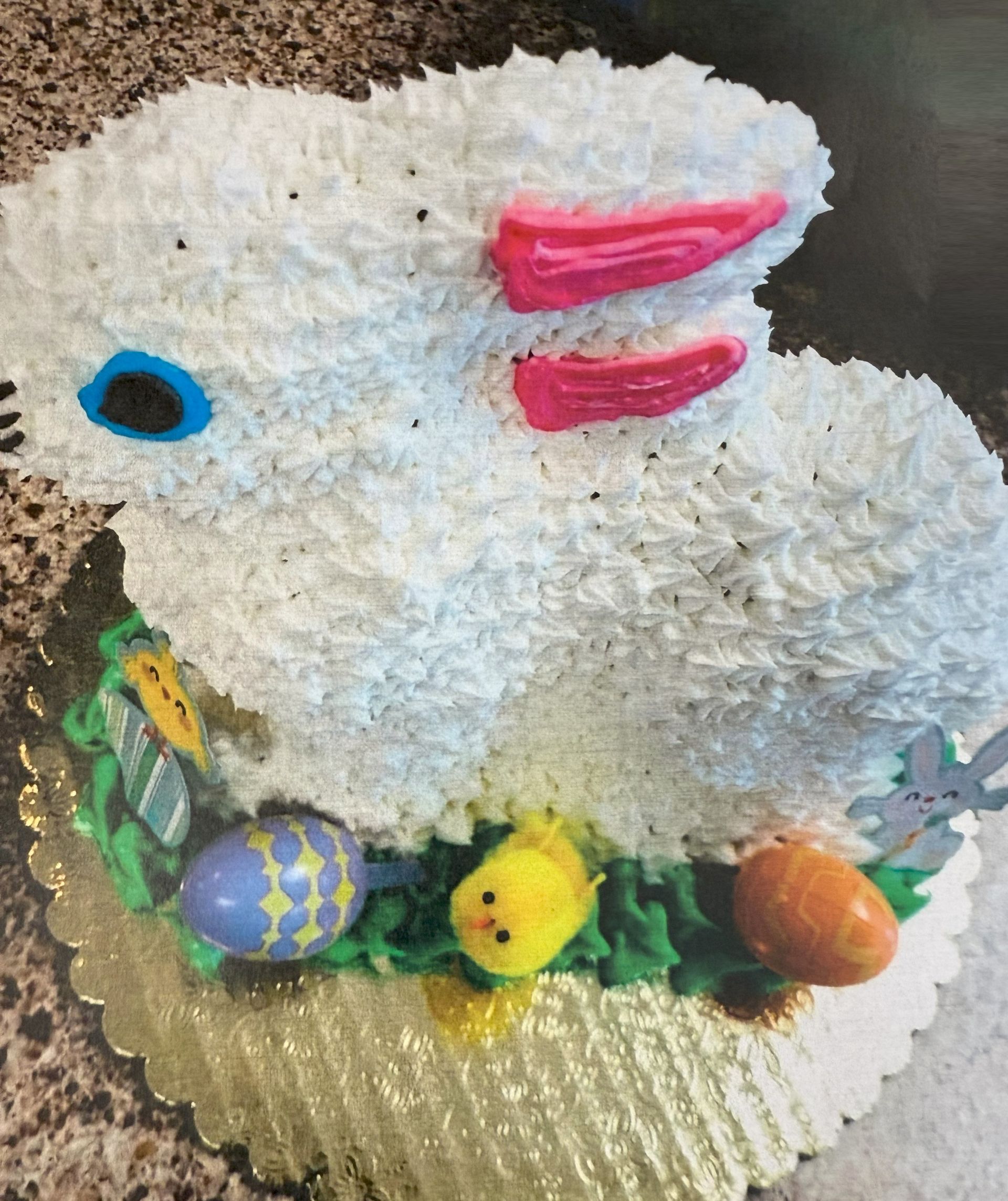 Easter bunny cake with white frosting, pink ears, blue eye, and decorated with eggs and a chick.