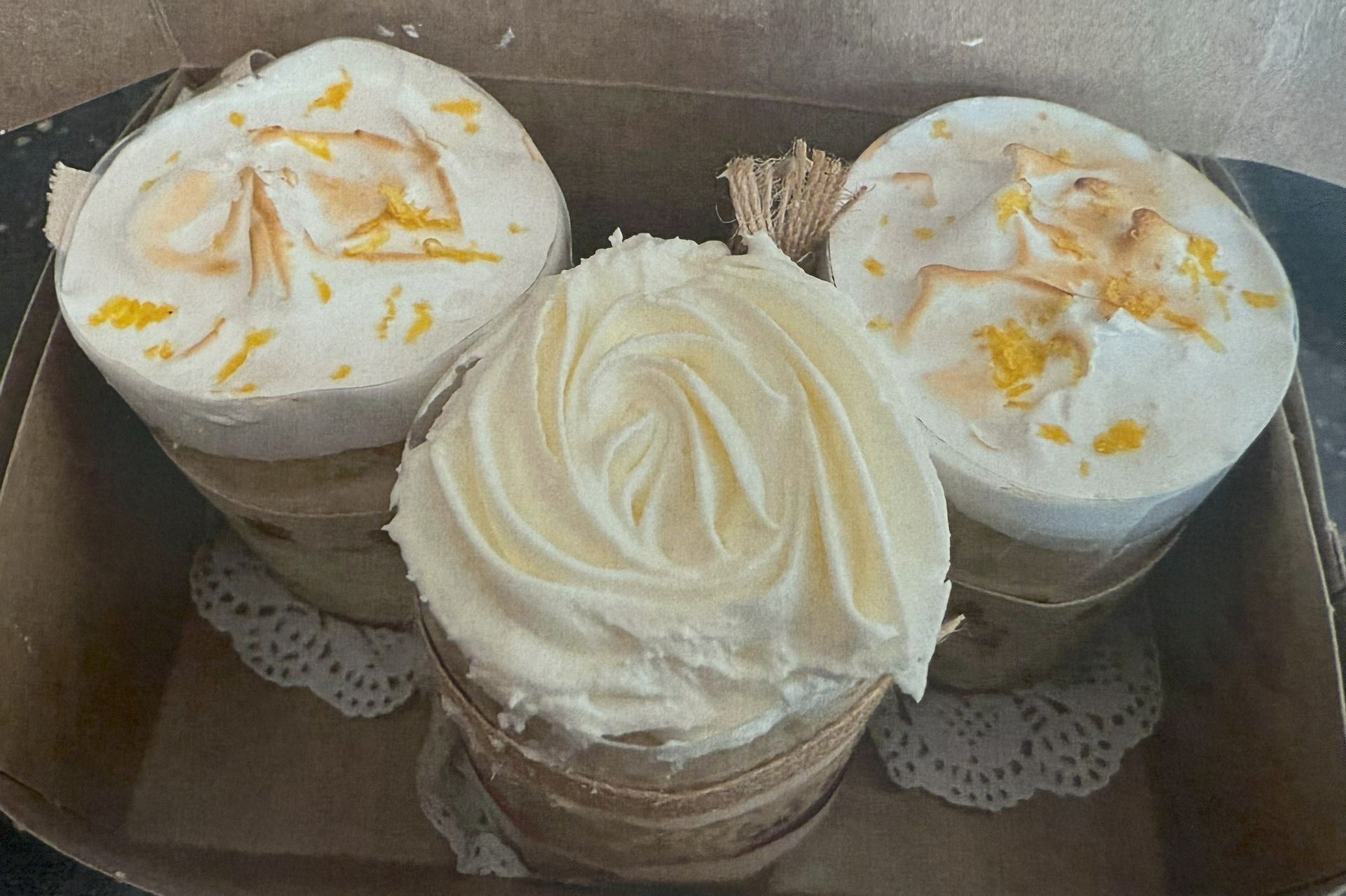 Three small round cakes in a box. White frosting, one rose-shaped. Decorated with petals.