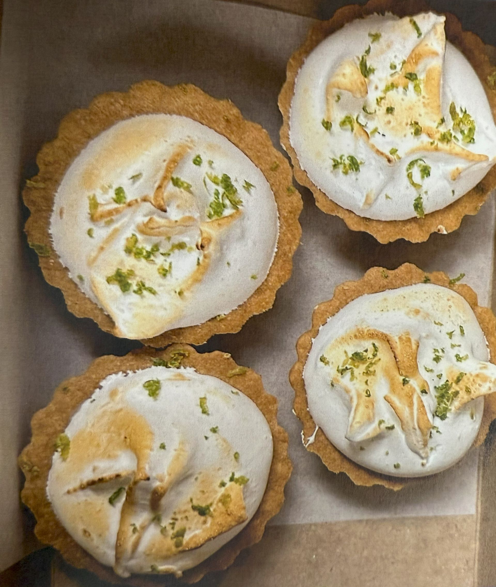 Four mini lemon meringue tarts in a box; browned crusts, white meringue, green zest.