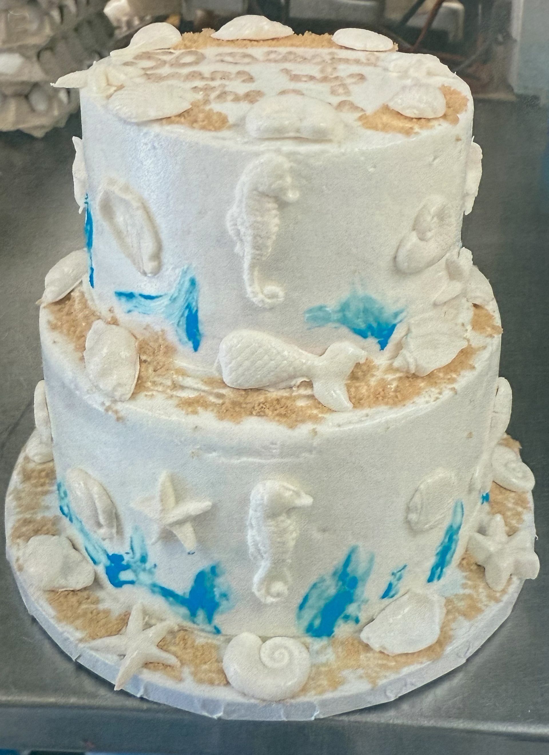 Two-tiered cake with beach theme: white frosting, seashells, seahorses, blue accents, and sandy accents.