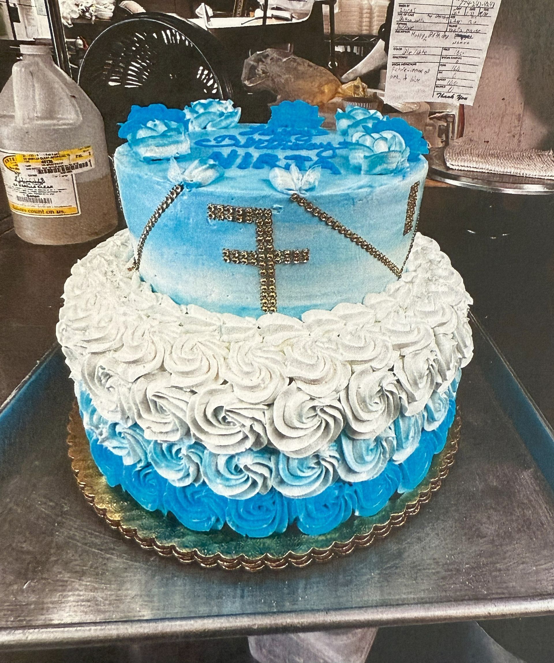 Two-tiered cake with white and blue frosting, decorated with a cross and flowers.