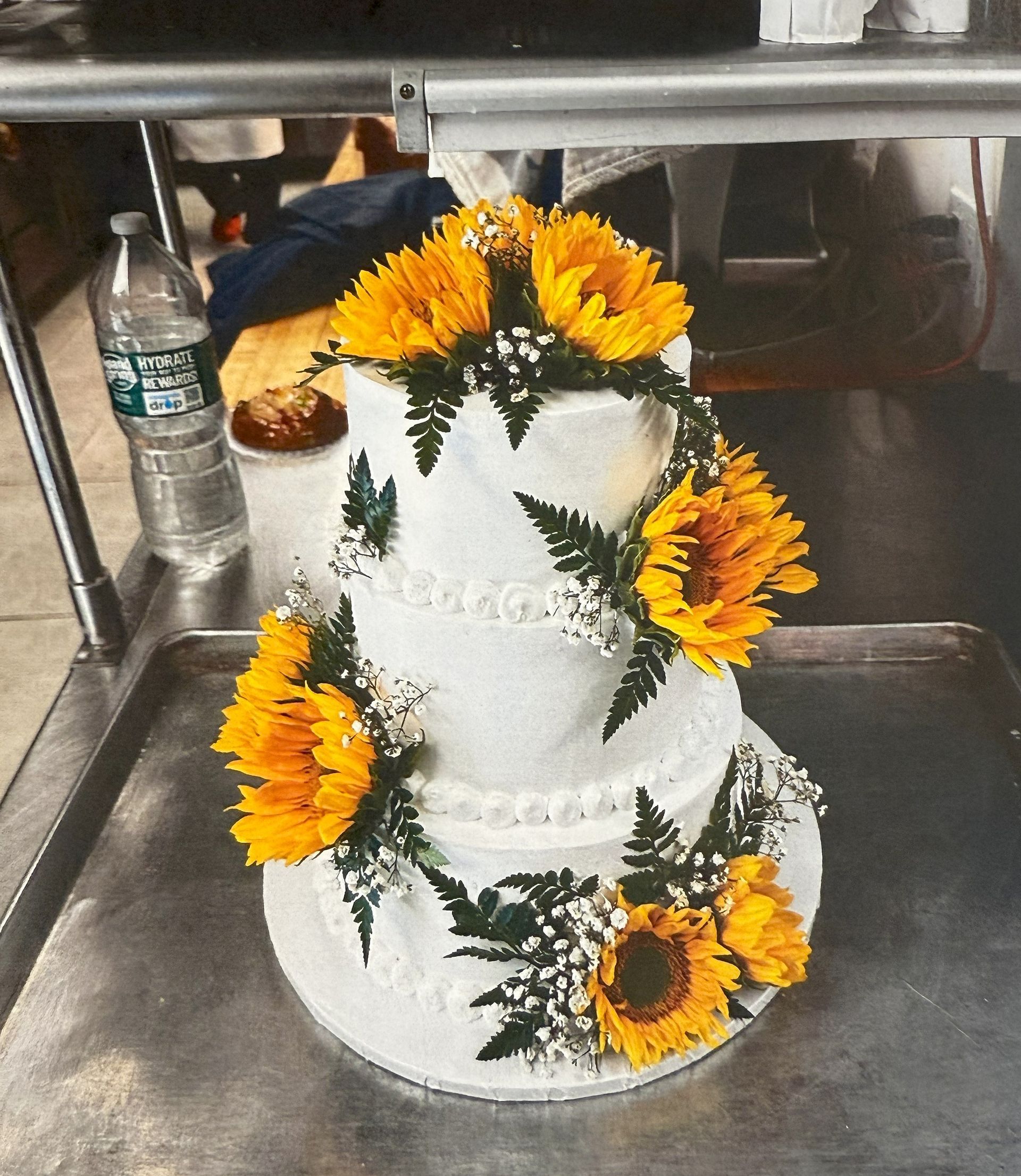 Three-tiered white cake decorated with yellow sunflowers and green ferns.