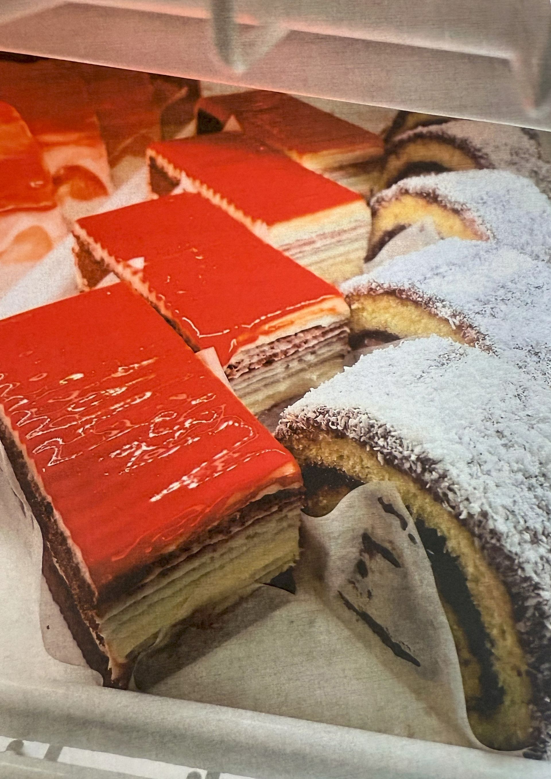 Display case filled with layered pastries: red-glazed rectangular cakes and snow-dusted rolls.