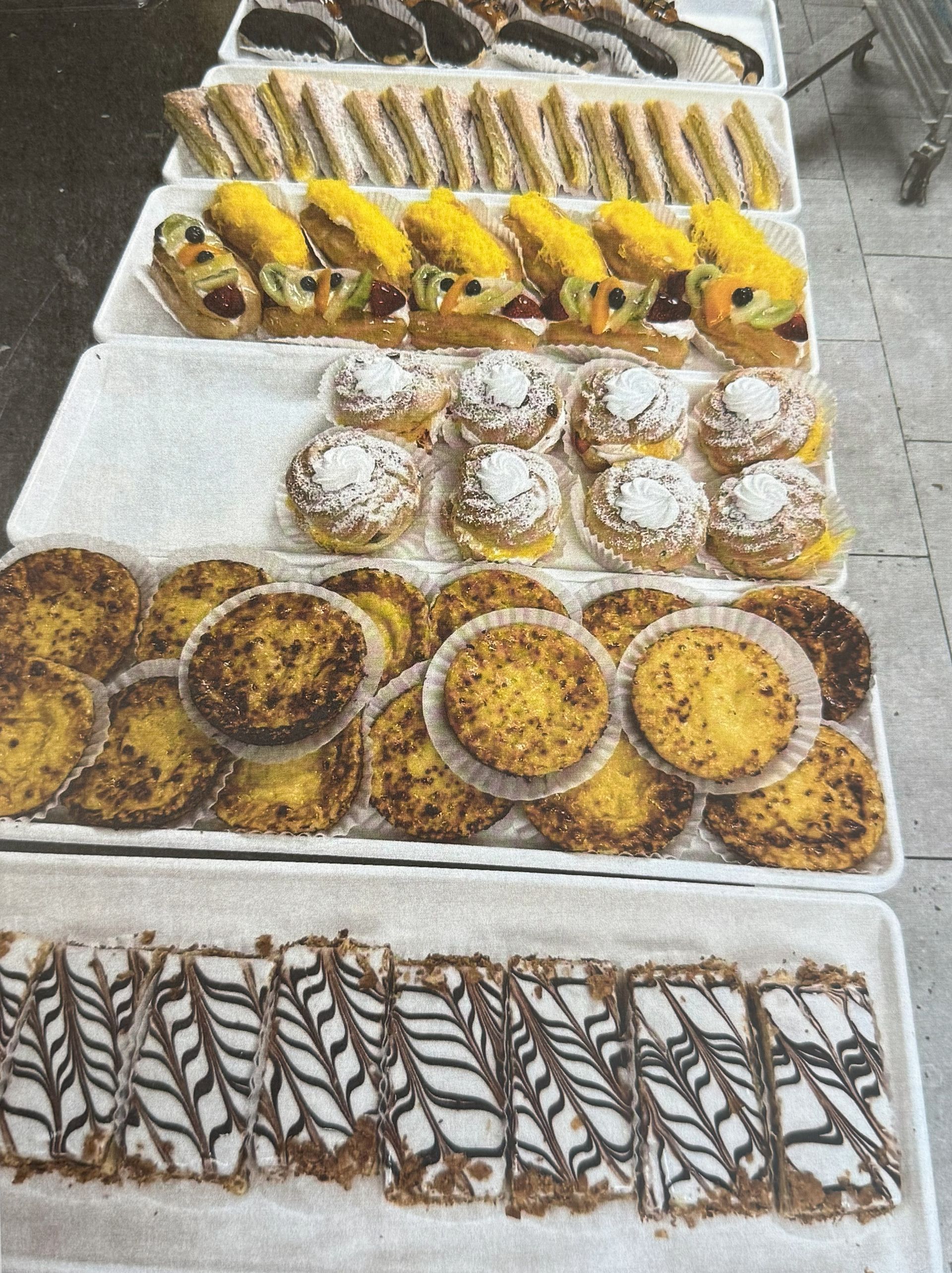Variety of pastries displayed on trays, including sandwiches, tarts, and danishes.