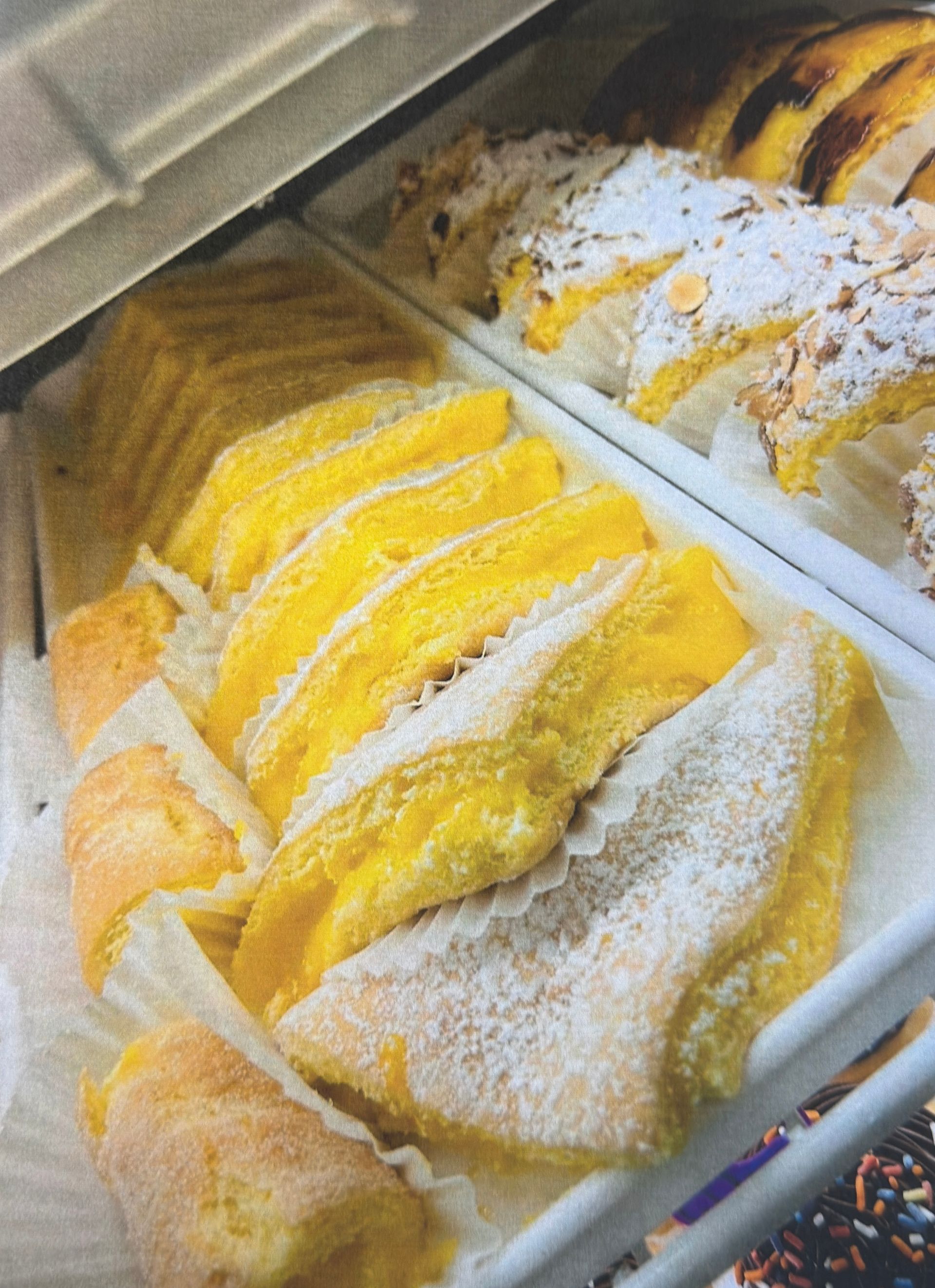 Yellow pastries in a bakery display case, including layered sponge cakes and rolled desserts, dusted with powdered sugar.