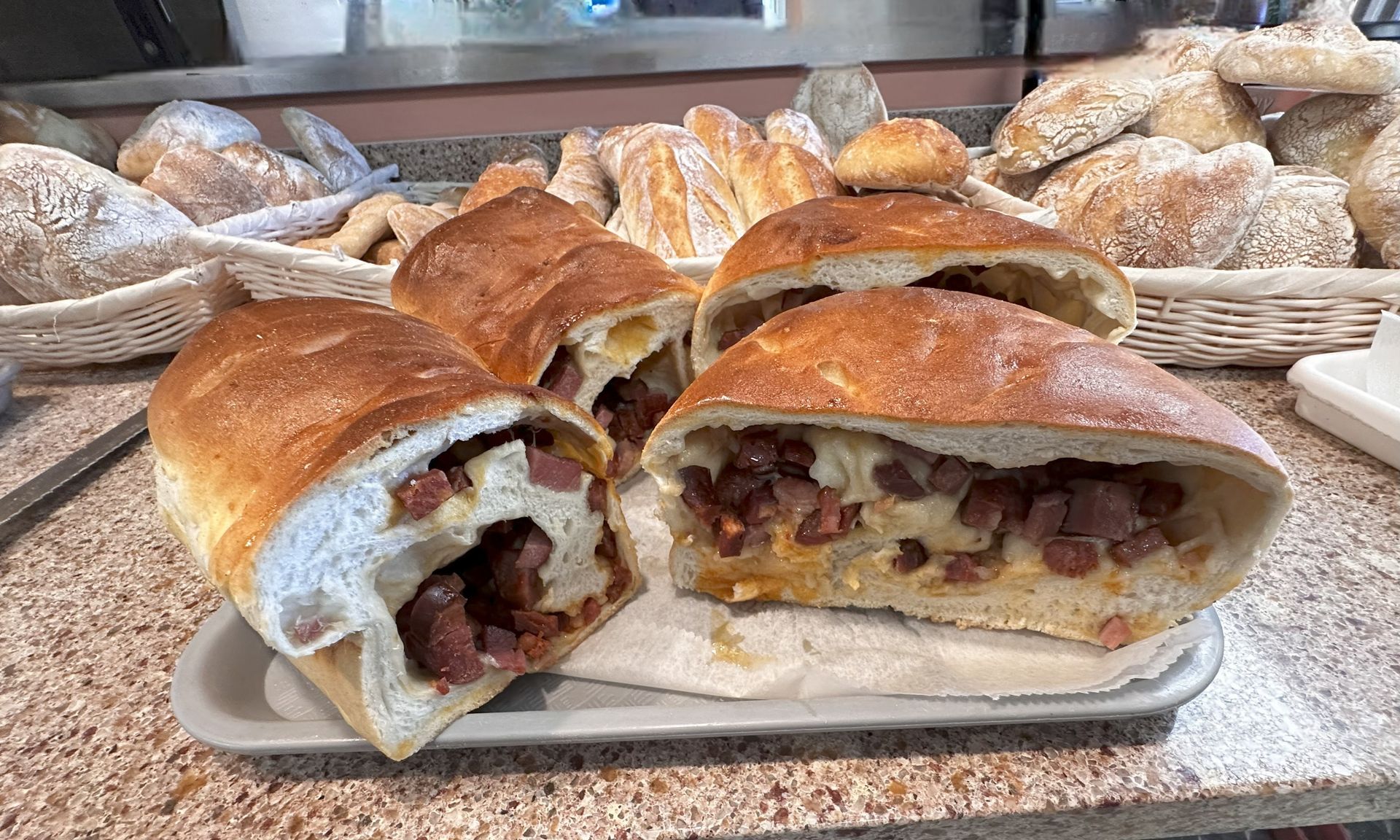 Bread loaves filled with meat, displayed on a tray. Other breads visible in the background.