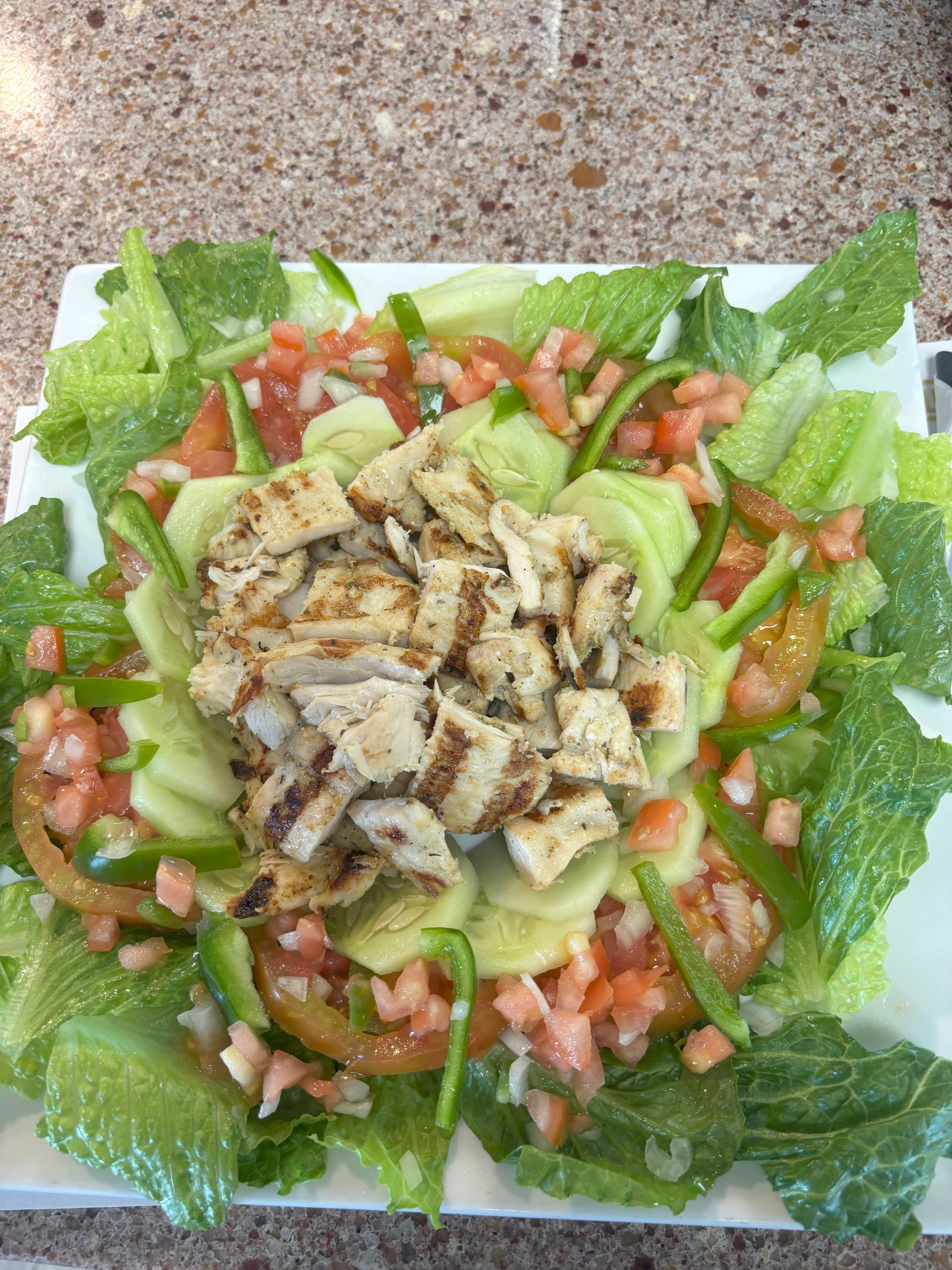 Grilled chicken salad with romaine lettuce, tomatoes, cucumber, and green peppers on a white plate.