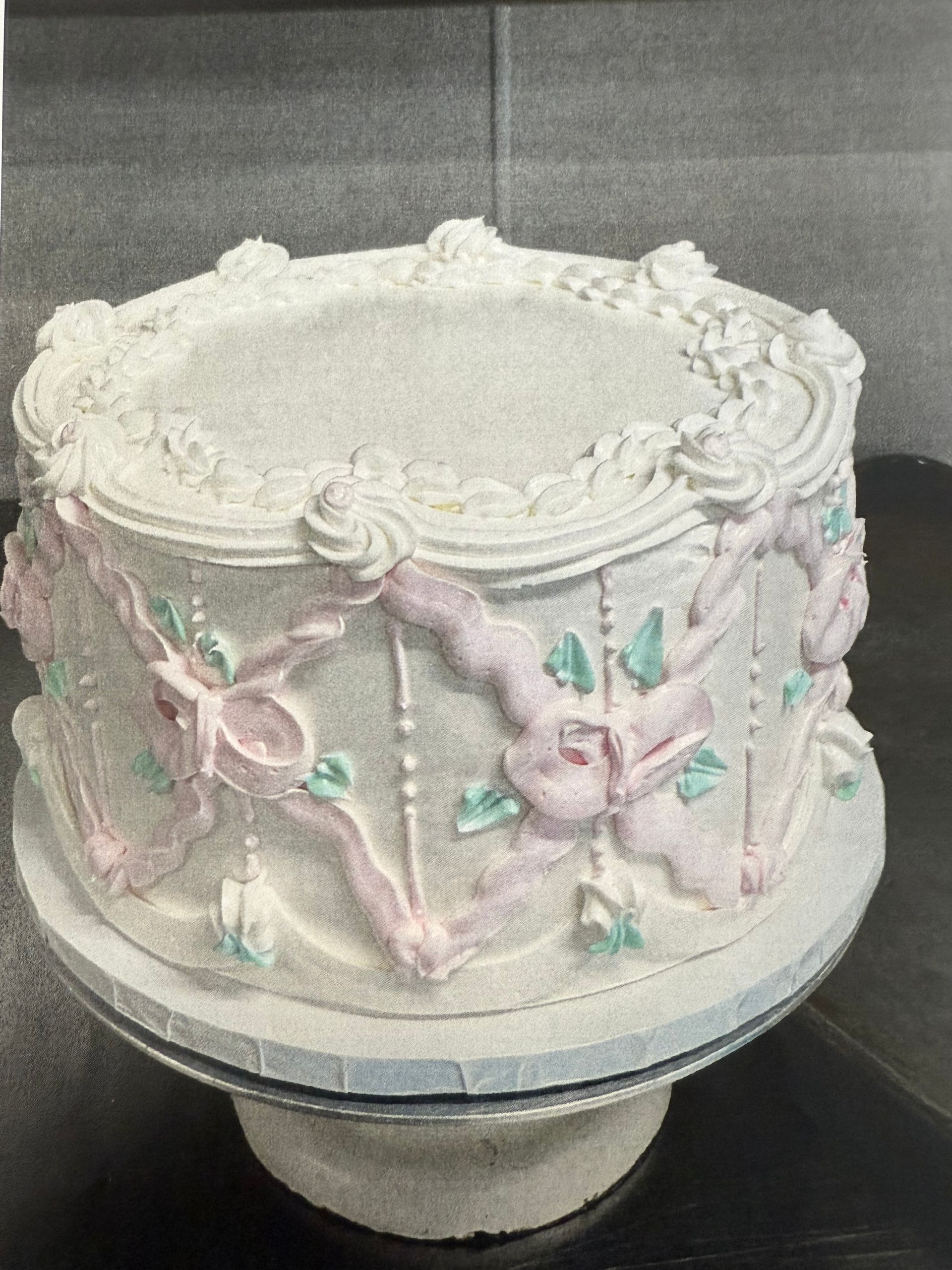 White cake with pink ribbons and green accents, on a silver cake stand.