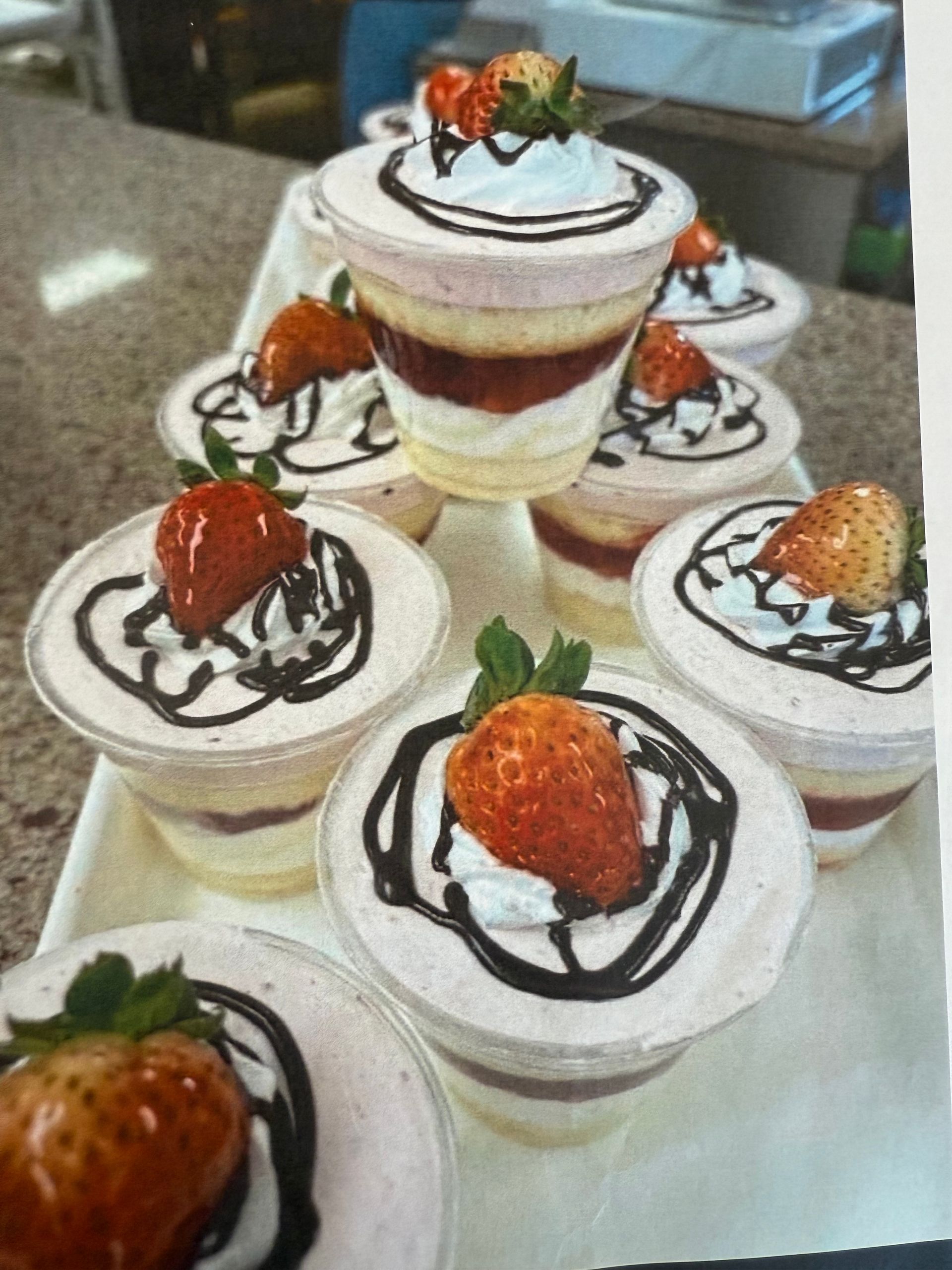 Strawberry parfaits in clear cups, topped with whipped cream, chocolate, and a whole strawberry on a tray.
