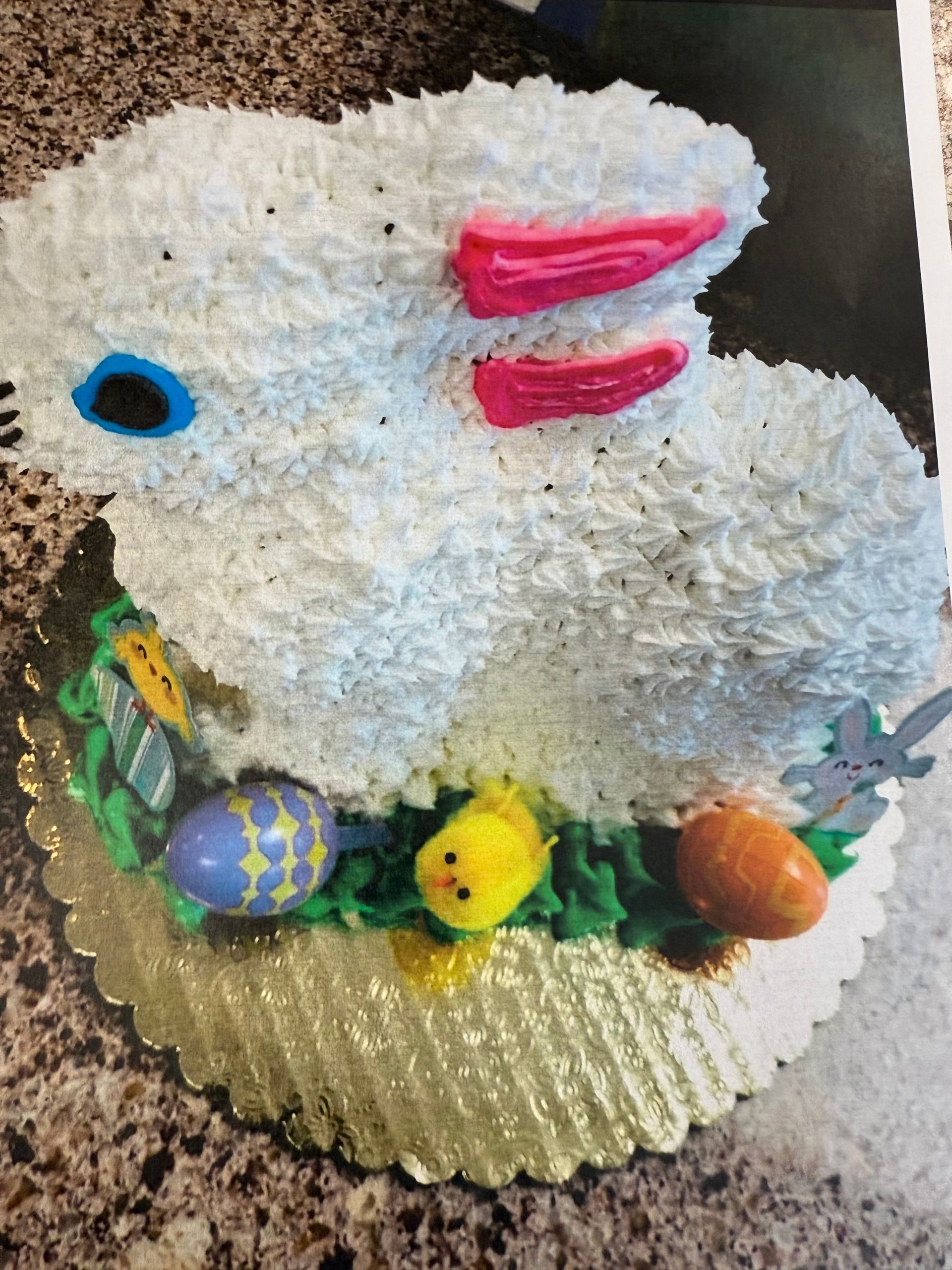 Easter bunny cake, white frosting with pink ears, blue eye, decorated with eggs and chick on a gold cake board.