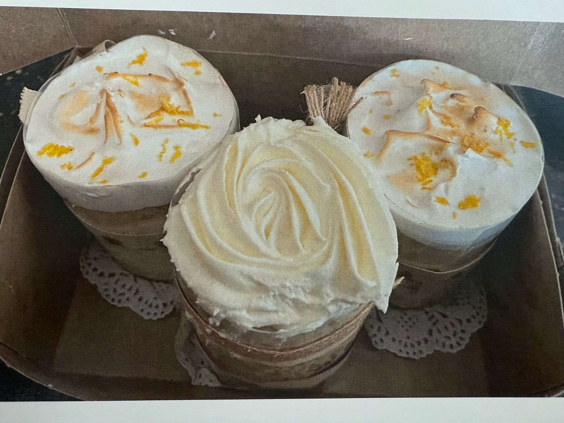 Three round, decorated cupcakes in a box. Two have floral toppings, the third has swirled frosting.