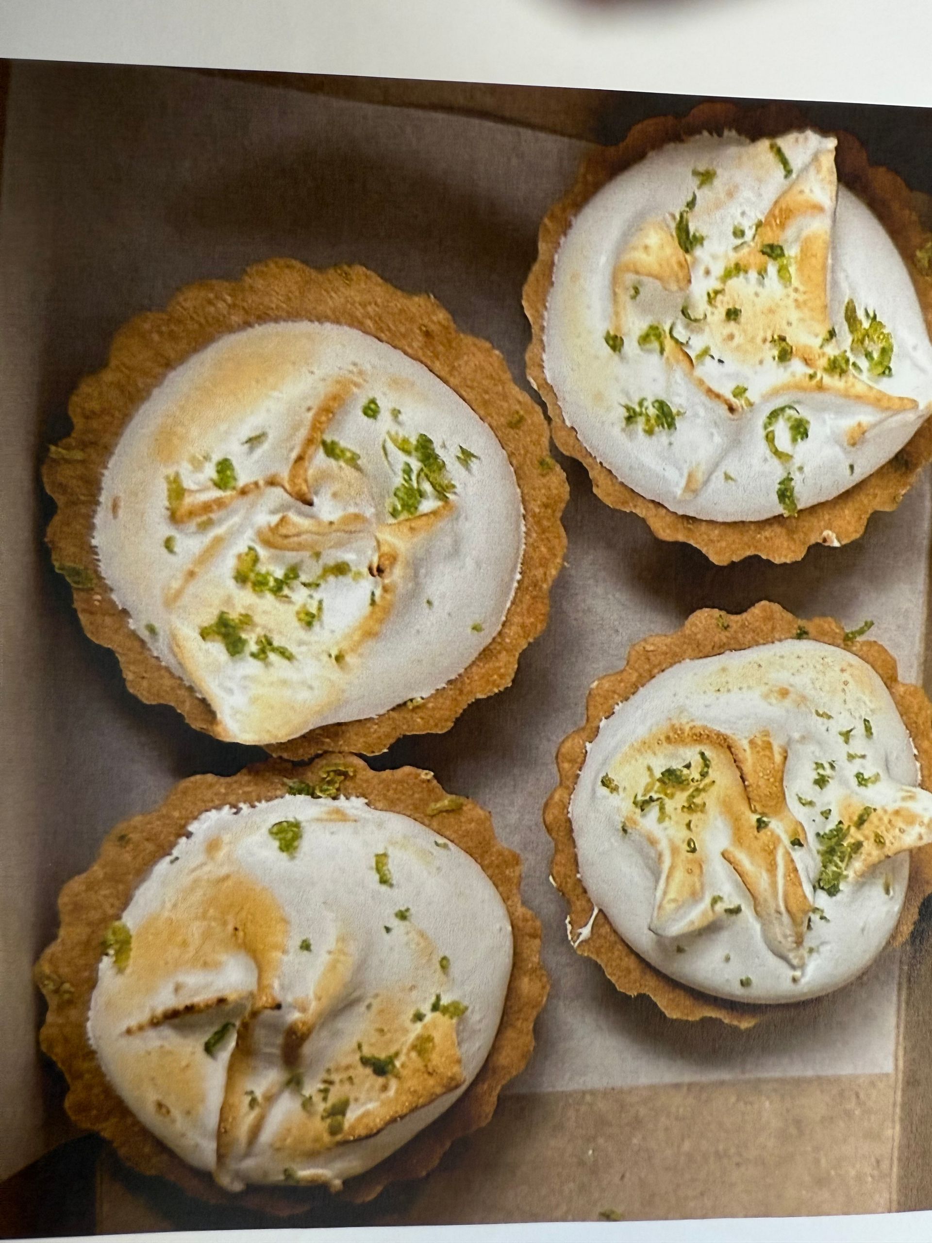 Four mini lemon meringue tarts in a box; golden crusts, white meringue peaks, and lime zest.