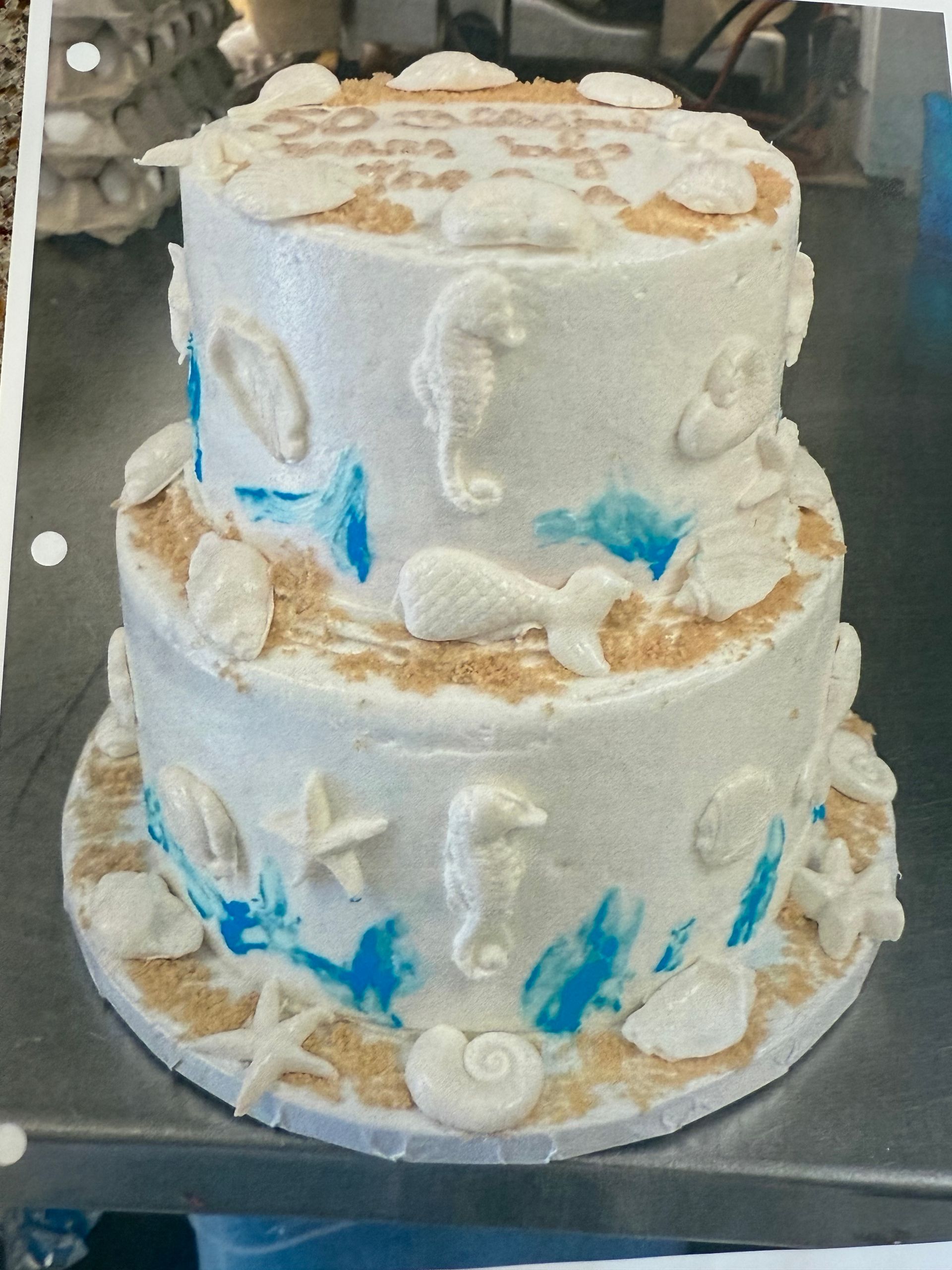 Two-tiered beach-themed cake with shells, seahorses, and blue wave accents, sitting on a metal surface.