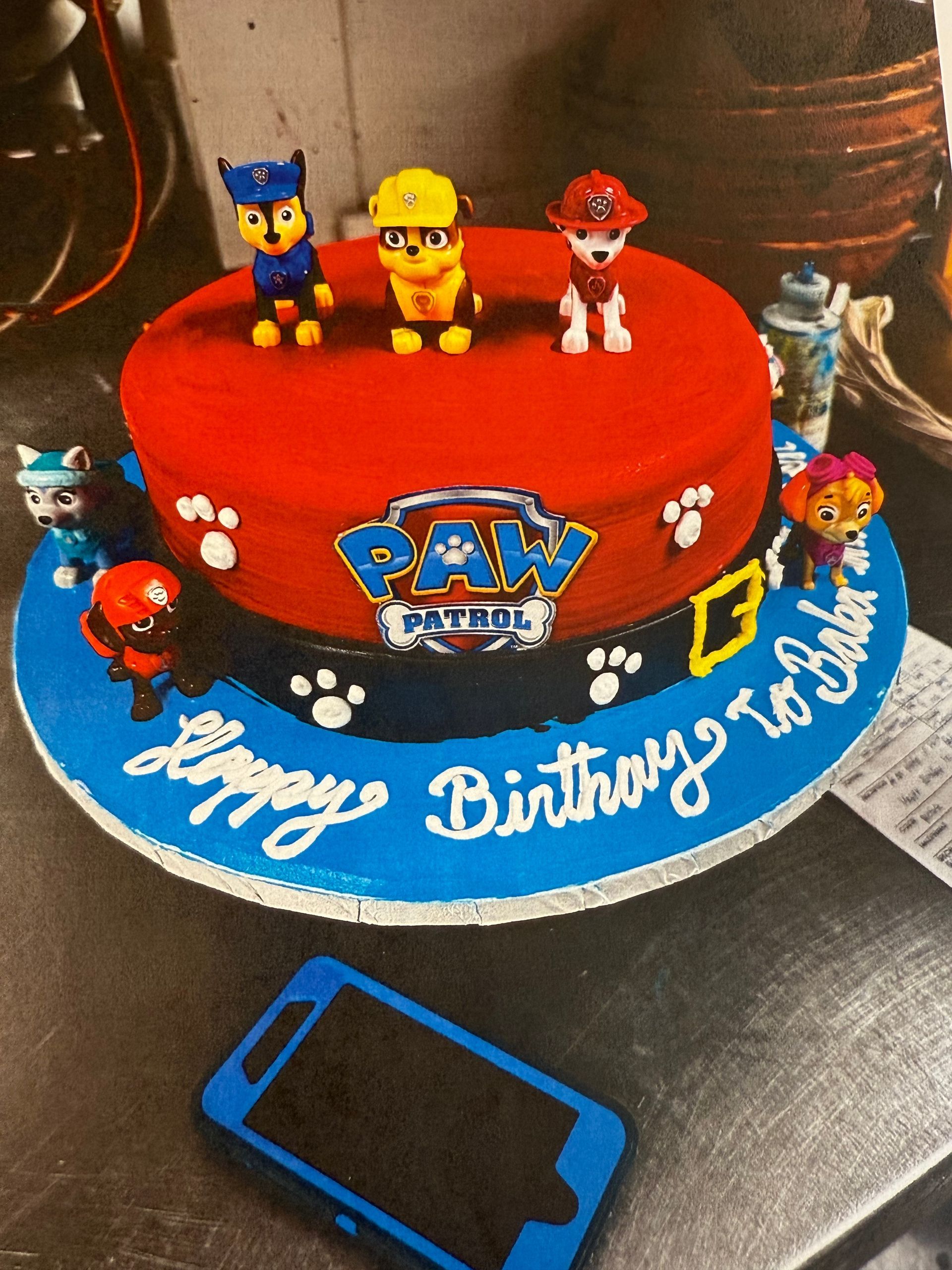 Paw Patrol birthday cake with figurines, red frosting, blue base, black strap, and 