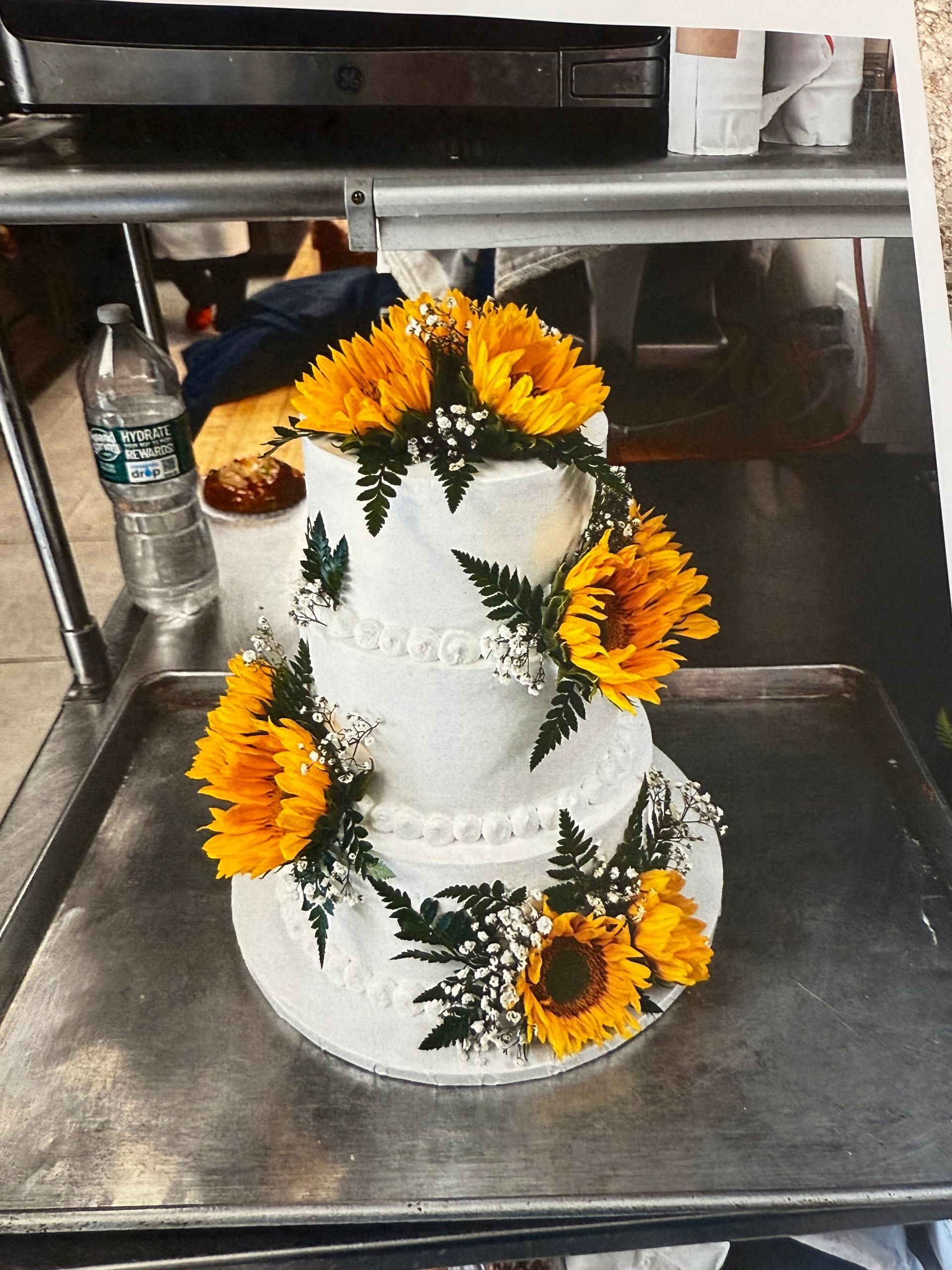 Three-tiered white cake decorated with yellow sunflowers and greenery on a metal tray.