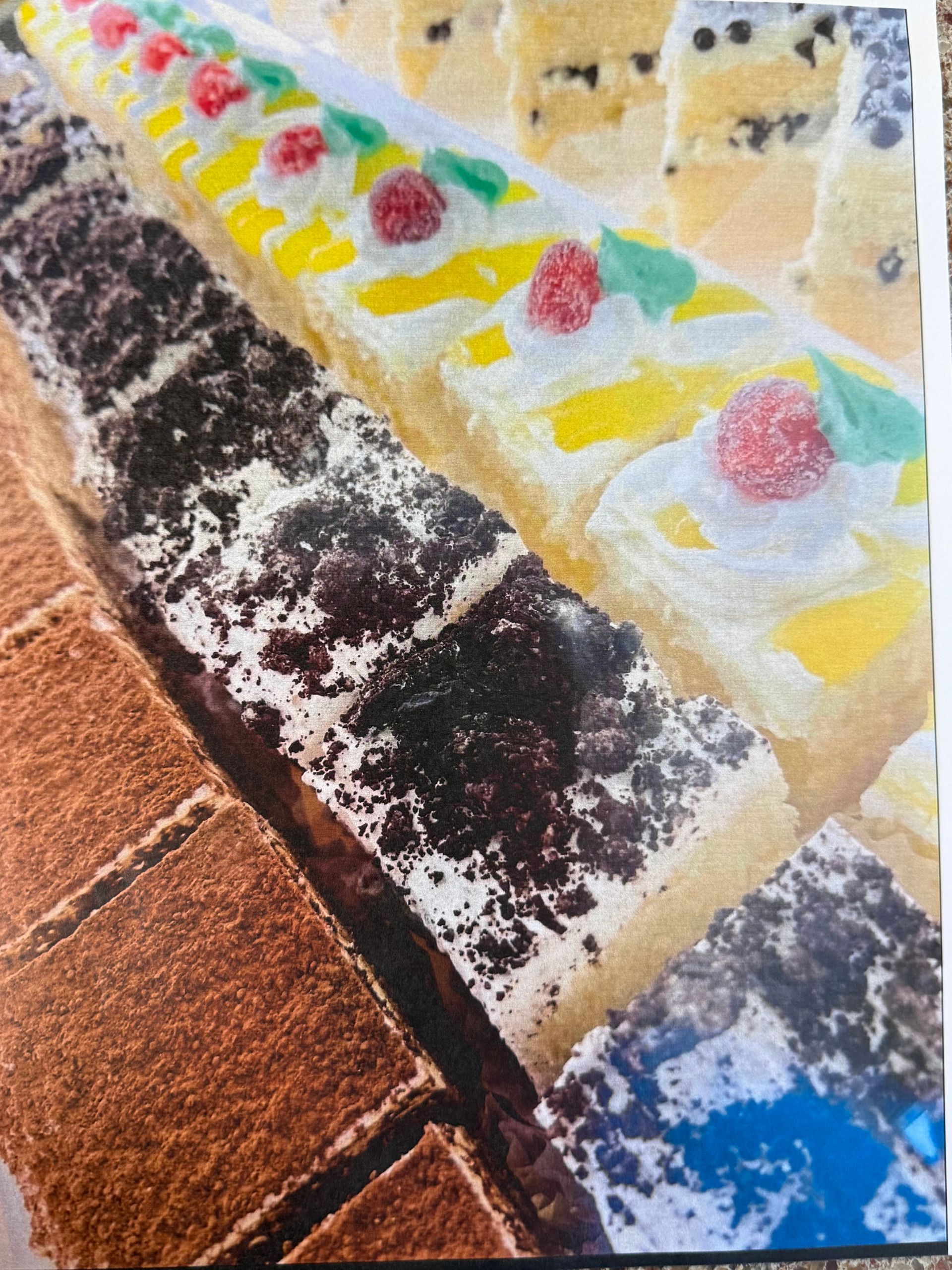 Rows of colorful, rectangular pastries: chocolate, yellow with red berries, and blue dusted with white powder.