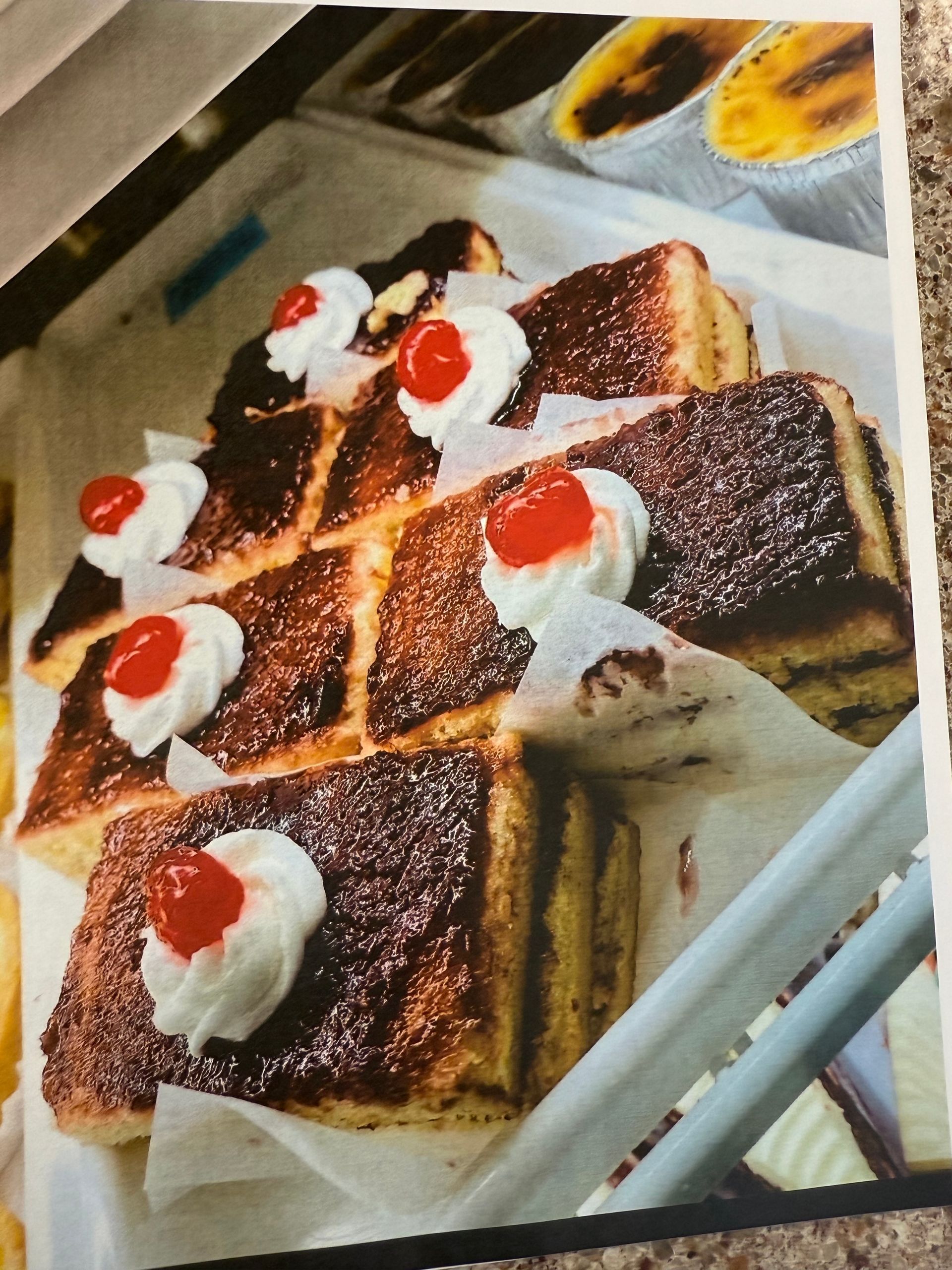 Cakes with brown tops, layered sides, whipped cream, and cherries on top in a display case.