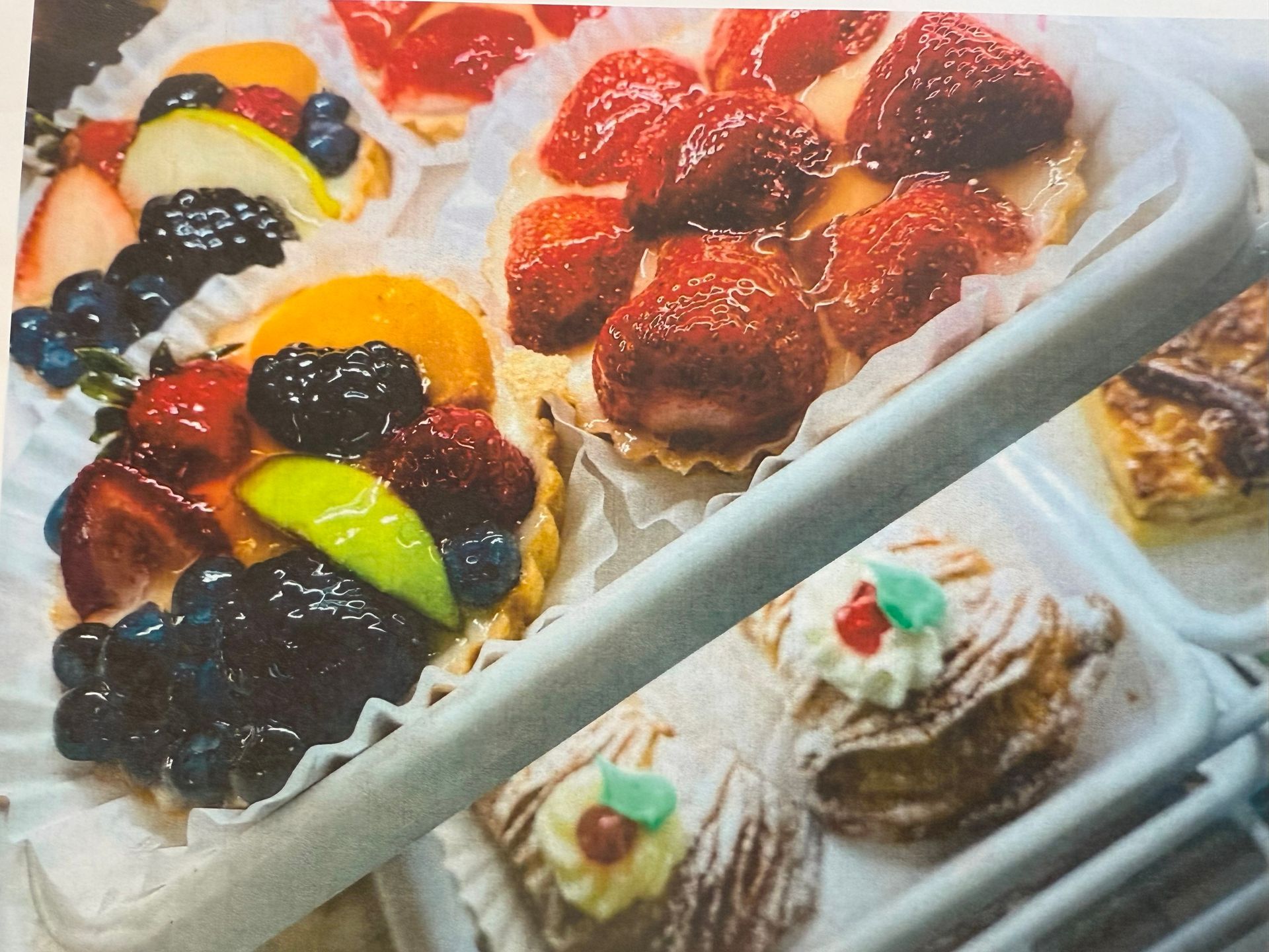 Fruit tarts with strawberries, blueberries, peaches, and pastries in a bakery display case.