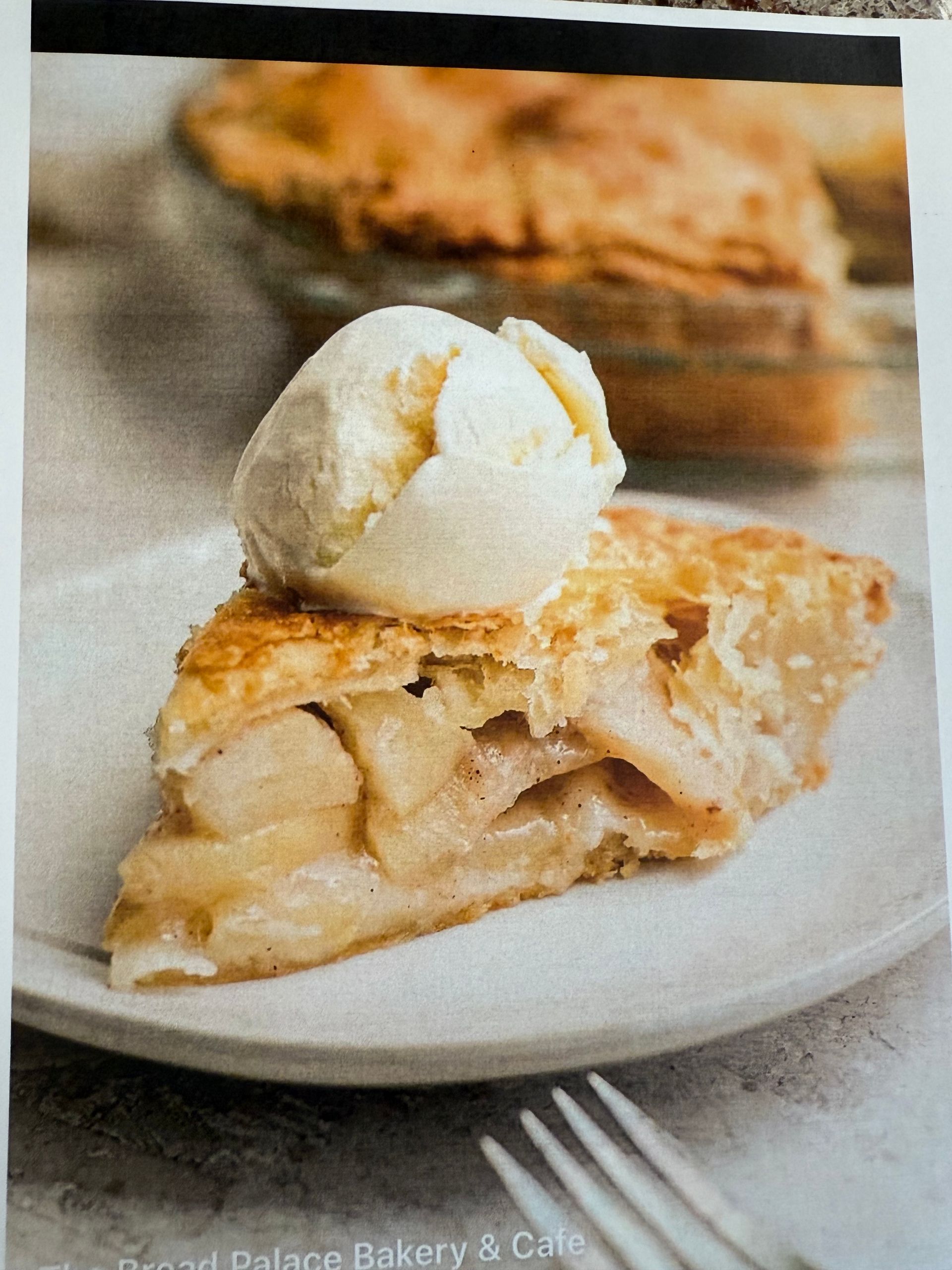 Slice of apple pie topped with vanilla ice cream on a plate, pie dish in the background.