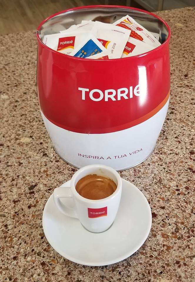 A coffee cup on saucer sits in front of a red and white Torrie sugar container filled with packets.