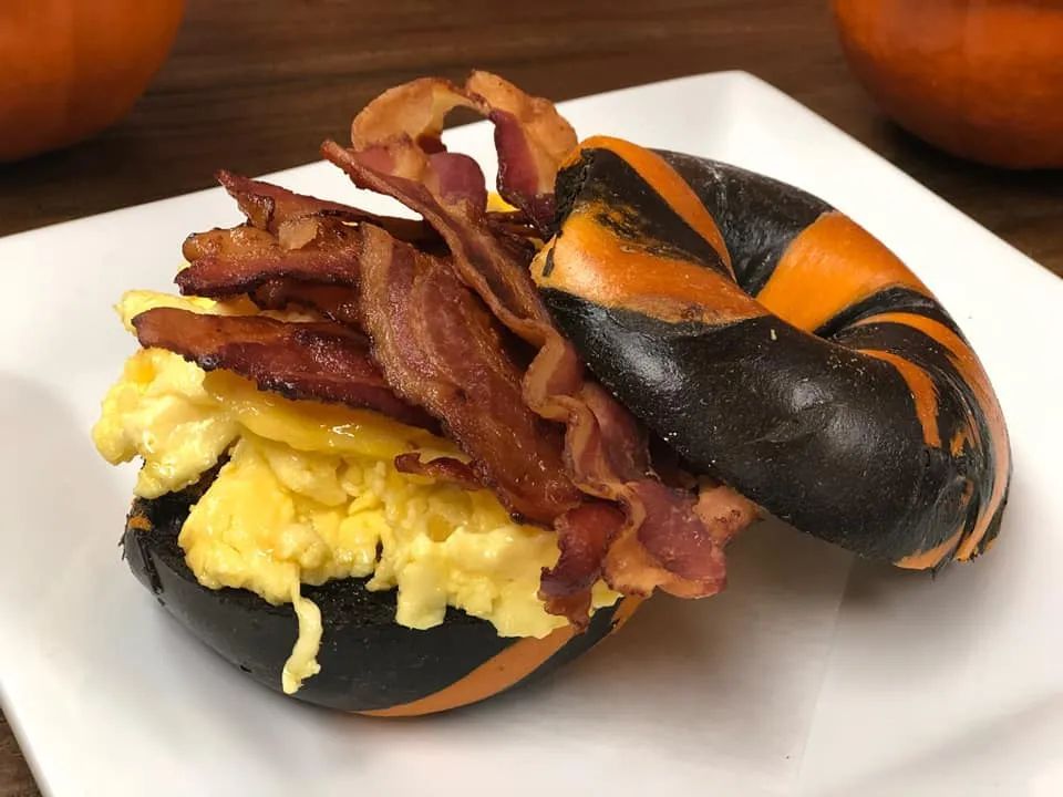 Halloween-themed bagel sandwich with scrambled eggs and bacon. Black and orange bagel on a white plate.