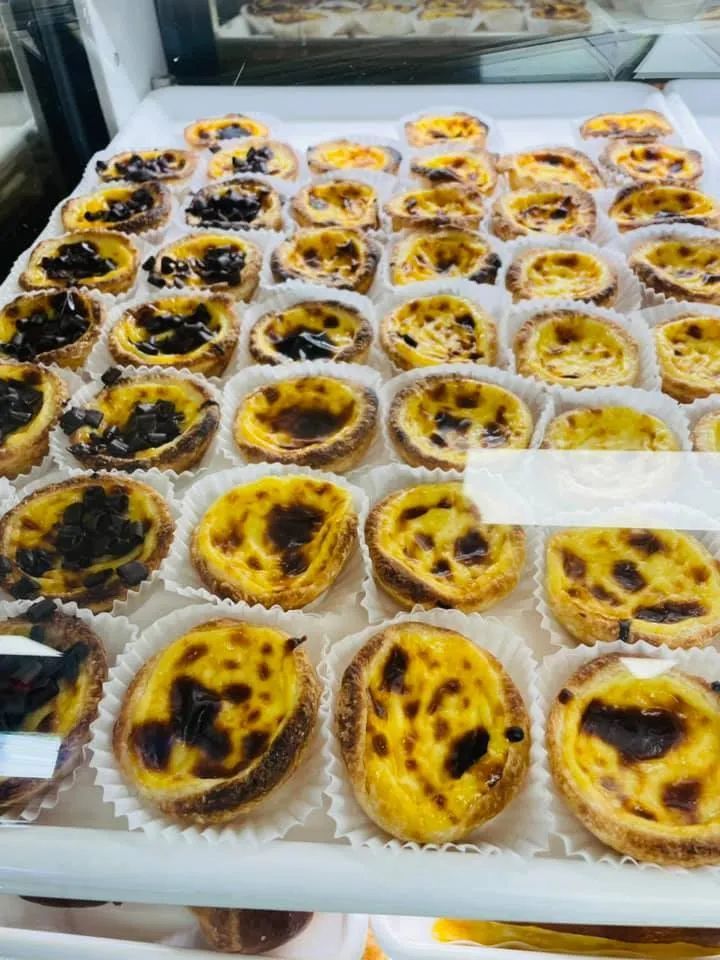 Rows of Portuguese custard tarts in paper cups, with caramelized tops, displayed in a bakery case.
