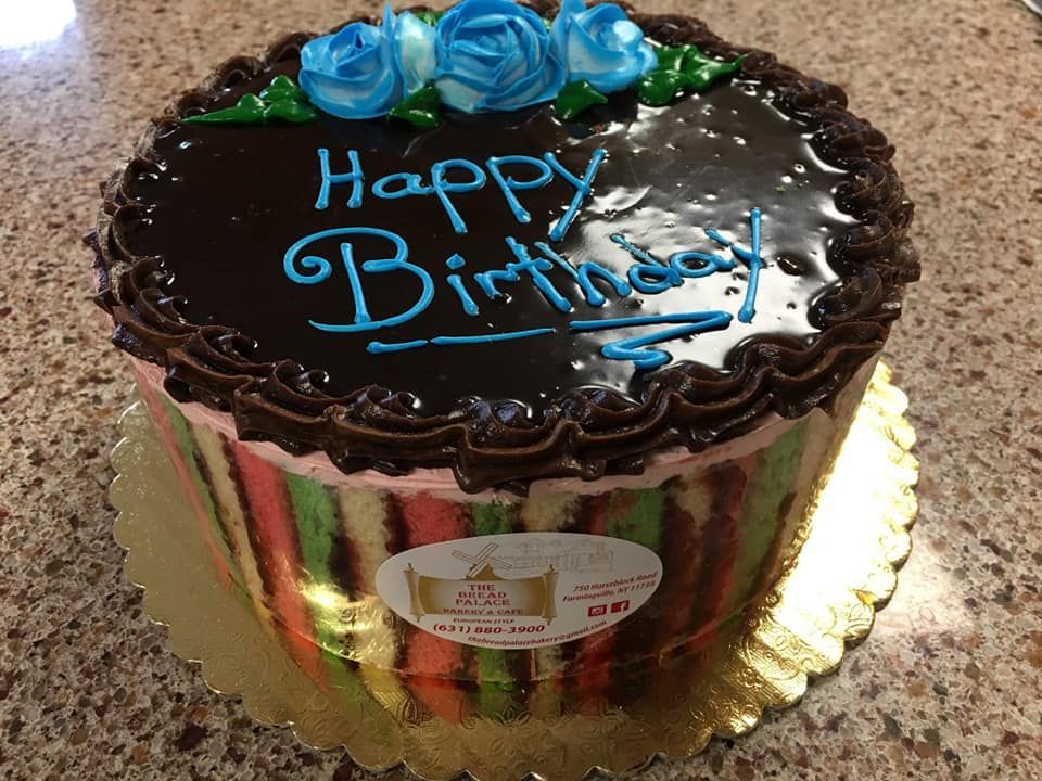 Birthday cake with colorful stripes, chocolate frosting, and blue roses on a gold stand.