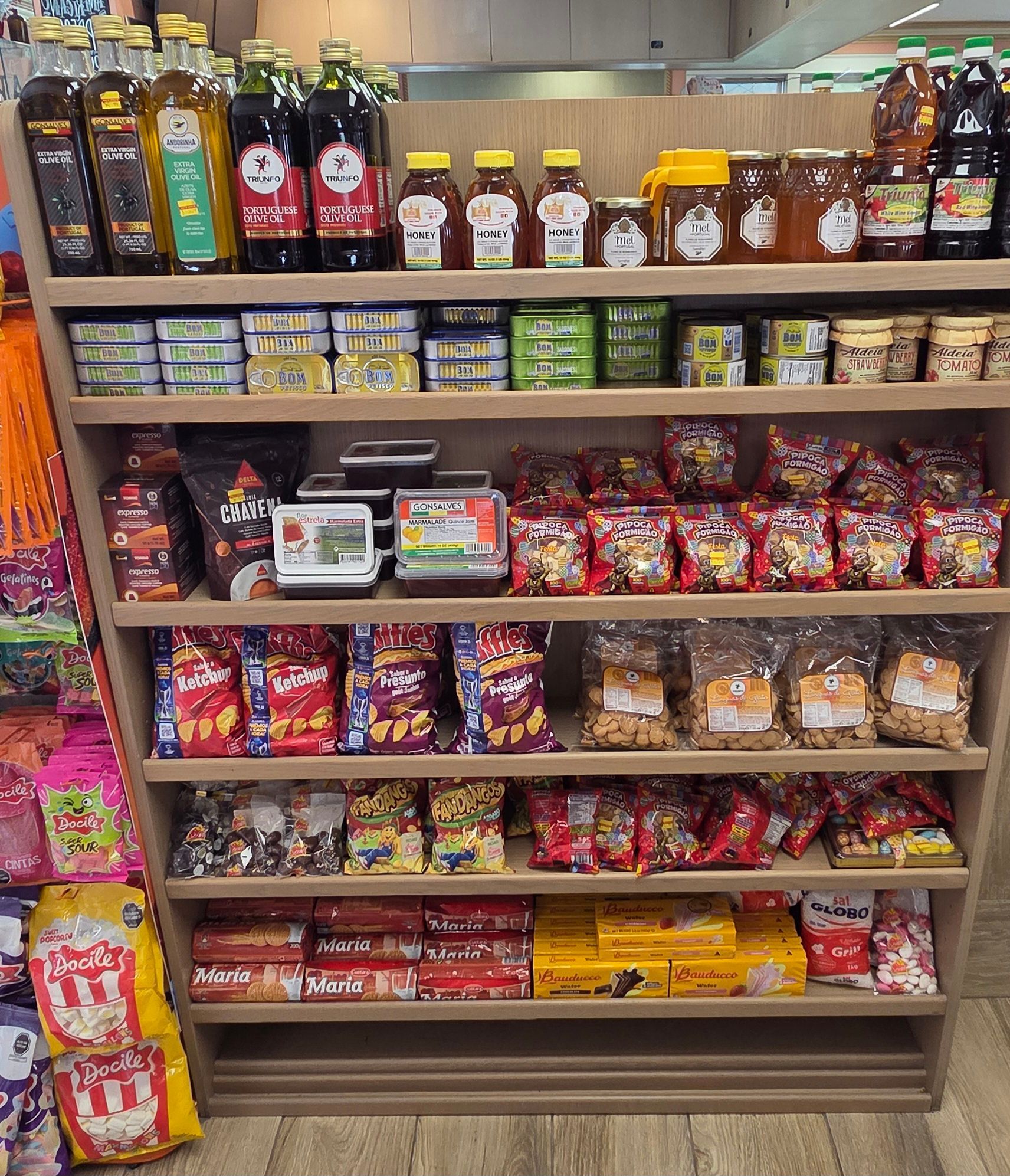 Wooden shelves stocked with food items, including oil bottles, honey jars, snacks, and condiments.