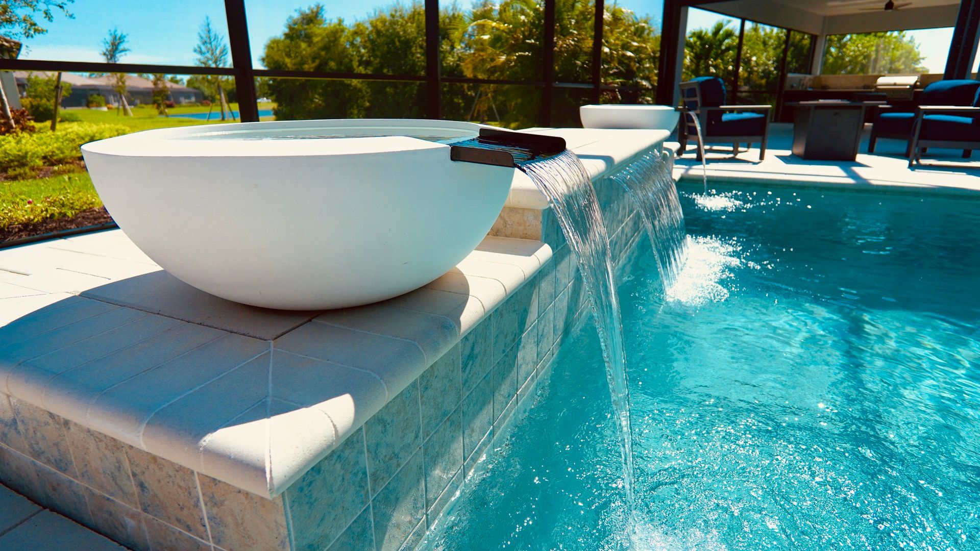 White bowl fountain overflowing into a turquoise swimming pool. Sunny outdoor patio.
