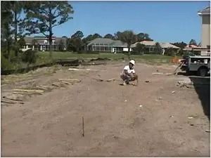 Construction worker on a dirt lot, leveling, with houses and blue sky in background.