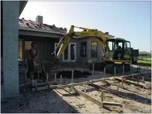Construction site: excavator near a house with a worker; forms laid for a concrete slab.