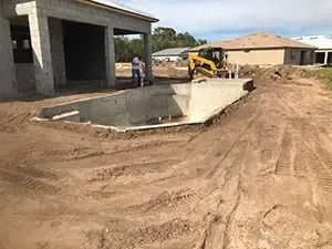 Pool under construction with backhoe and workers. Dirt surrounds the pool with a house in the background.