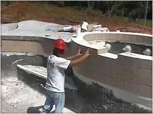 A construction worker in a hard hat tiling a swimming pool wall outdoors.