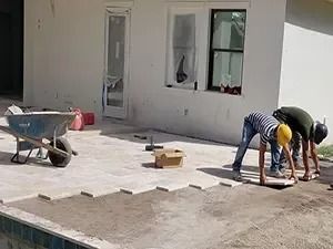 Two workers laying paving stones on a patio next to a pool. A wheelbarrow and tools are nearby.