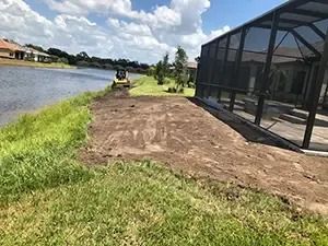 A lakeside yard with a pool screen, grass, and a cleared patch of dirt where a small machine is working.